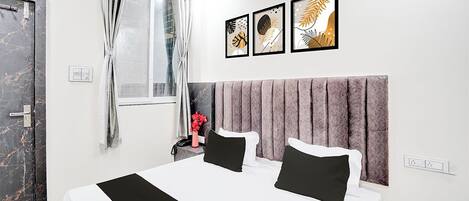 A hotel room with a large bed, a headboard, a window with curtains, and three framed artworks on the wall.
