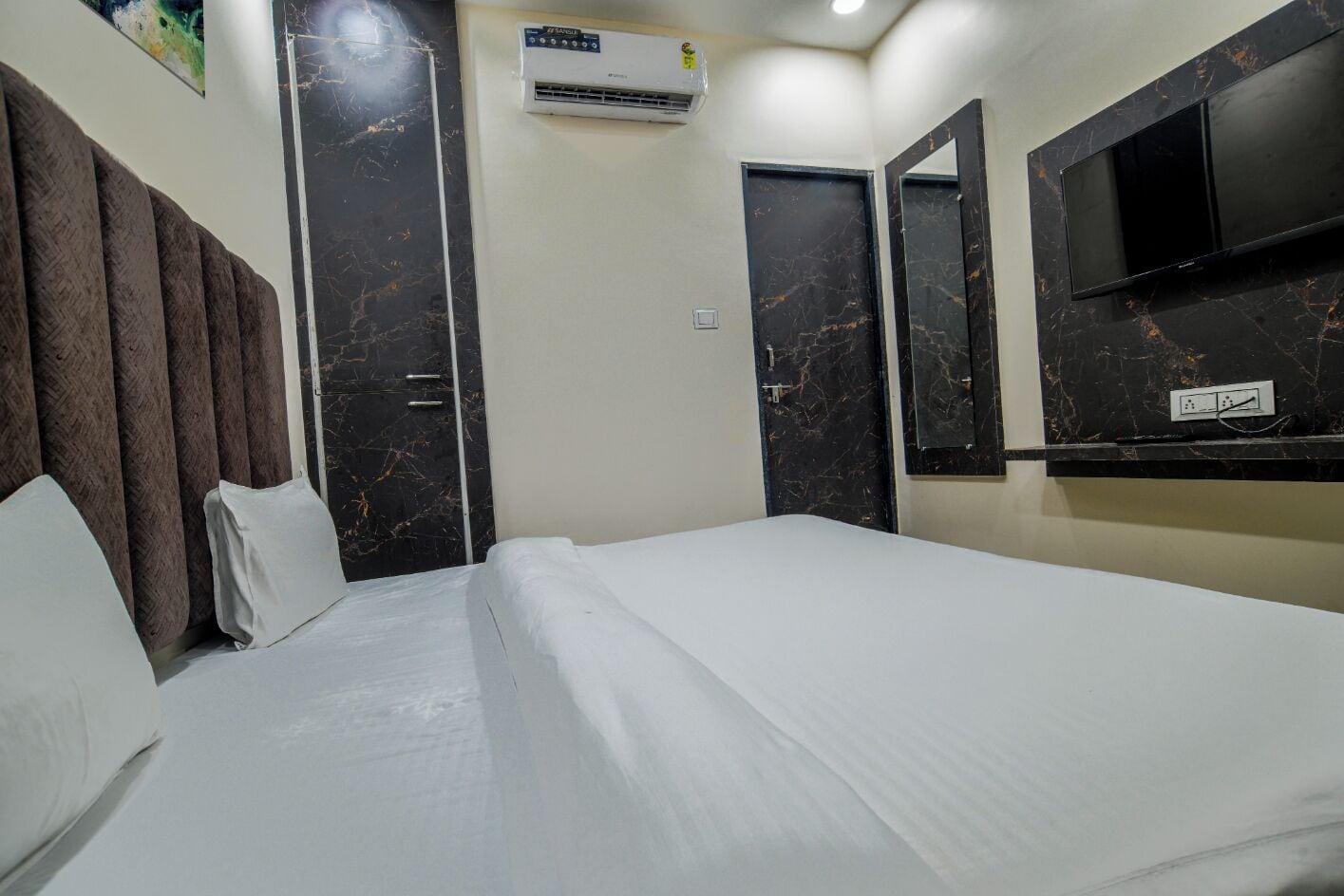 A hotel room with a bed, a flat-screen TV mounted on the wall, a wardrobe with a mirror, and an air conditioning unit.