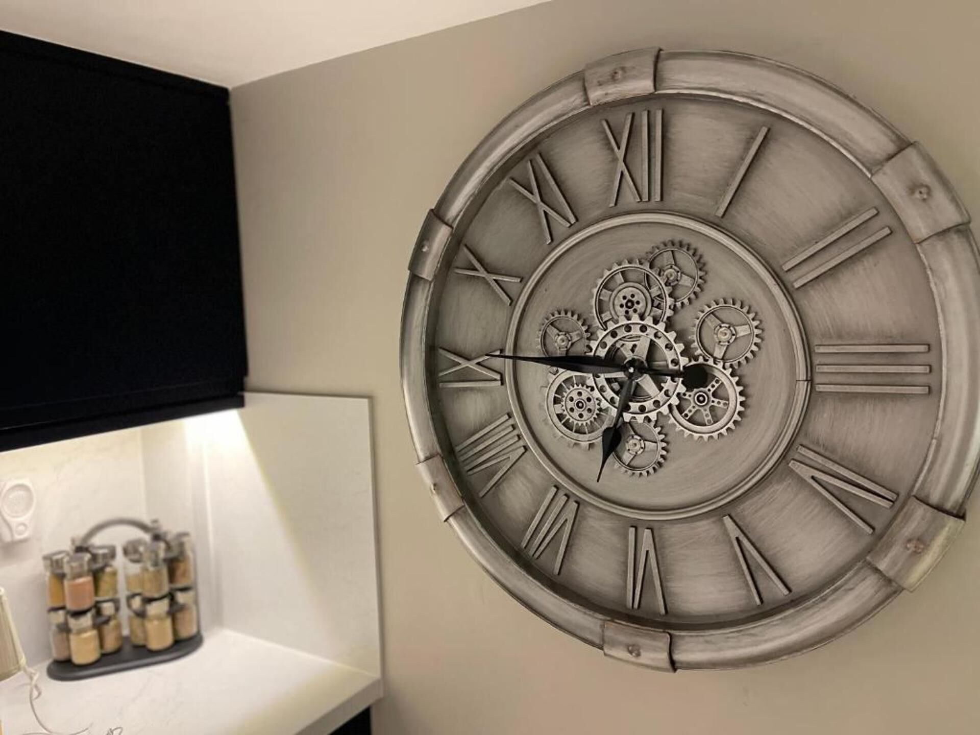 Un reloj de pared con números romanos y engranajes, situado en una habitación con un estante blanco y frascos.