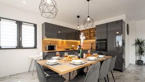 A modern dining area with a wooden table set for six, surrounded by grey upholstered chairs. A kitchen with dark cabinets and a microwave is visible in the background. - Awesome home in Jursici (Jursici)