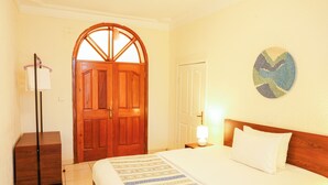 A hotel room with a wooden door, a bed with white bedding, a bedside table with a lamp, and a wall decoration. - Kalih Villa (Kigali)