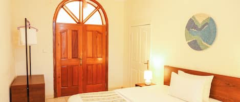 A hotel room with a wooden door, a bed with white bedding, a bedside table with a lamp, and a wall decoration.
