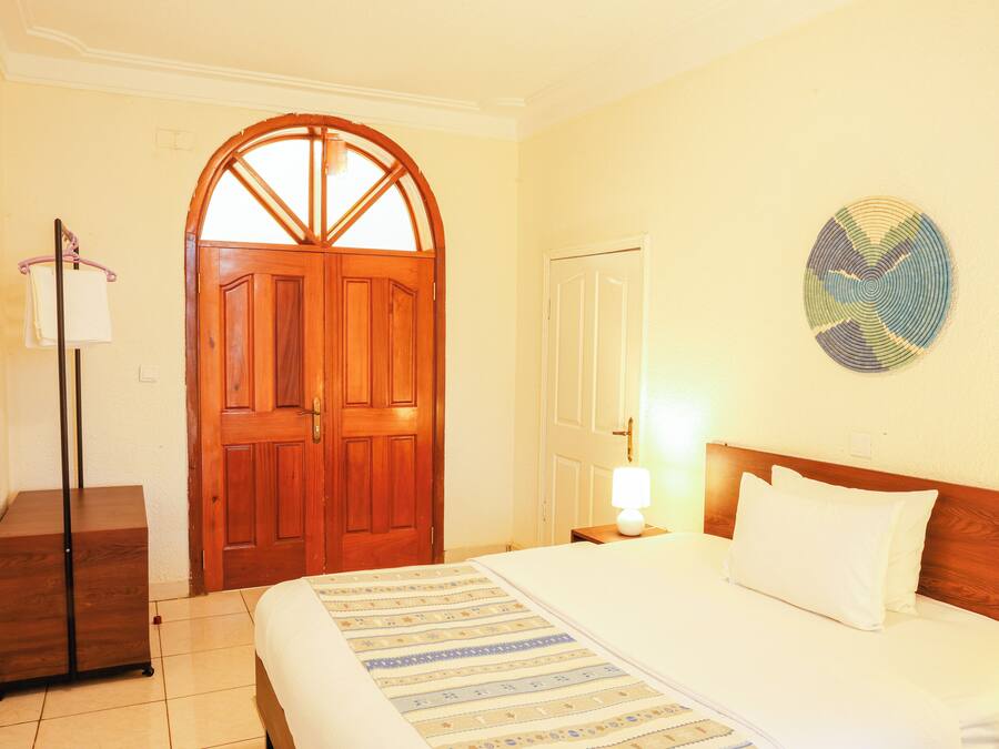 Habitación de hotel con puerta de madera, cama con ropa blanca, mesita de noche con lámpara y decoración en la pared.