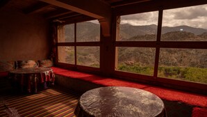 A room with a large window offering a mountain view, a round table with a patterned tablecloth, and a red carpeted area. - Gite Azoul (Beni Mellal)