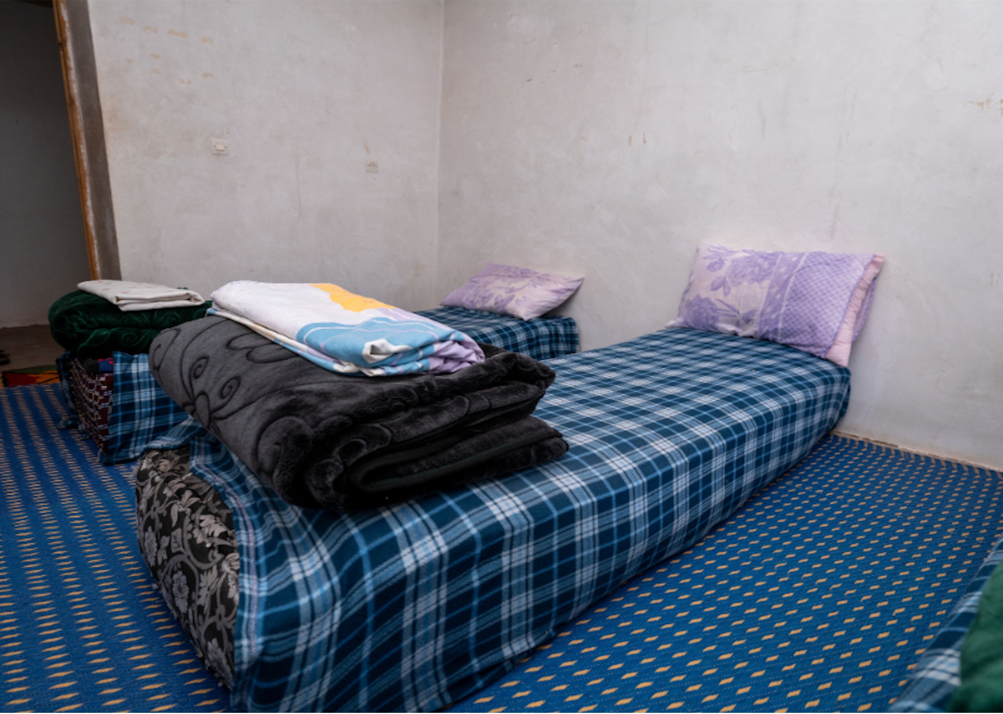 A small, single-bed room with a blue checkered bedspread, a purple pillow, and a folded blanket on the bed.