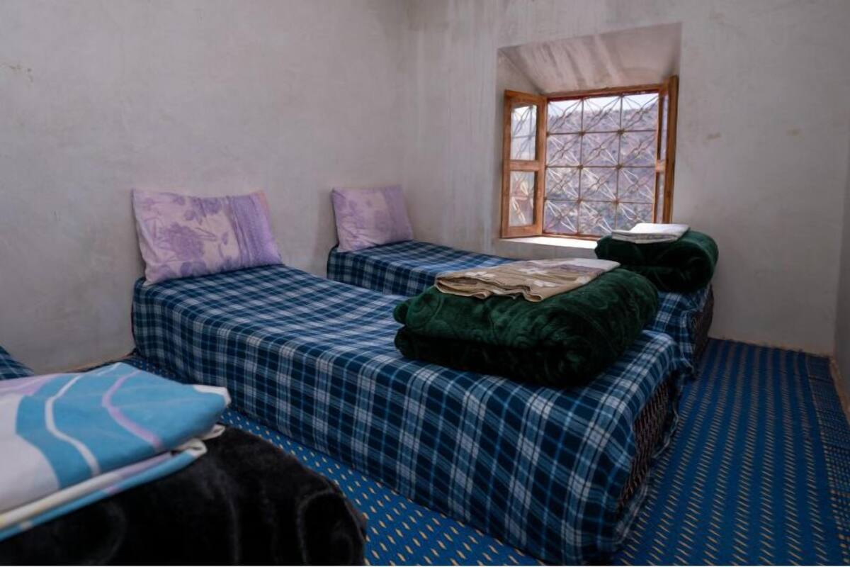 A small, sparsely furnished room with two beds, a window, and a plain wall.