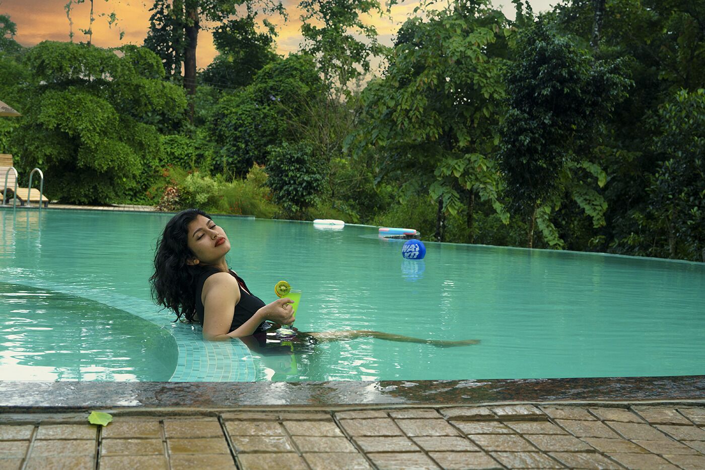 Una persona in piscina con un drink, immersa nel verde.