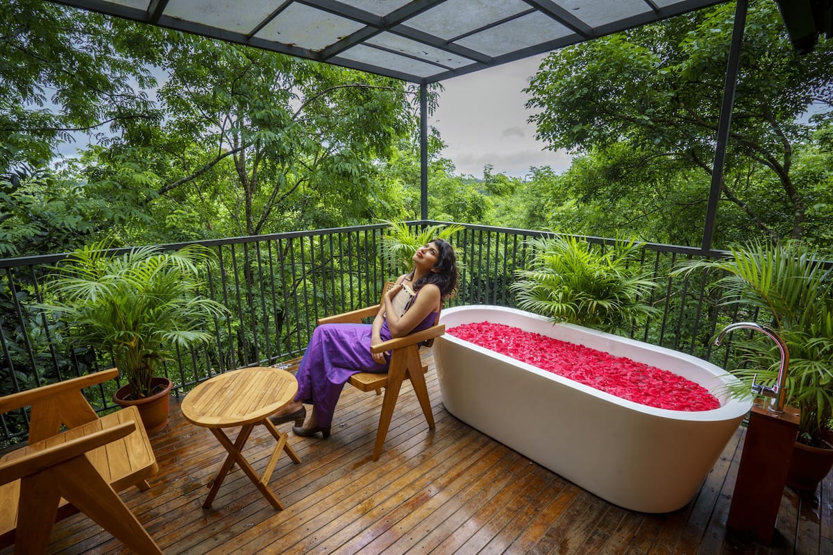 A woman sitting on a wooden chair by a white bathtub filled with red pebbles, surrounded by lush greenery on a wooden deck.