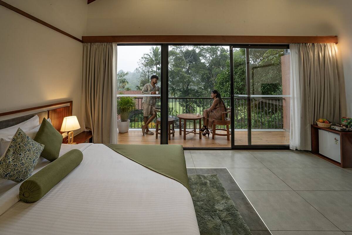 A hotel room with a bed, a nightstand, a lamp, a window with curtains, a balcony with a table and chairs, and a view of greenery outside.