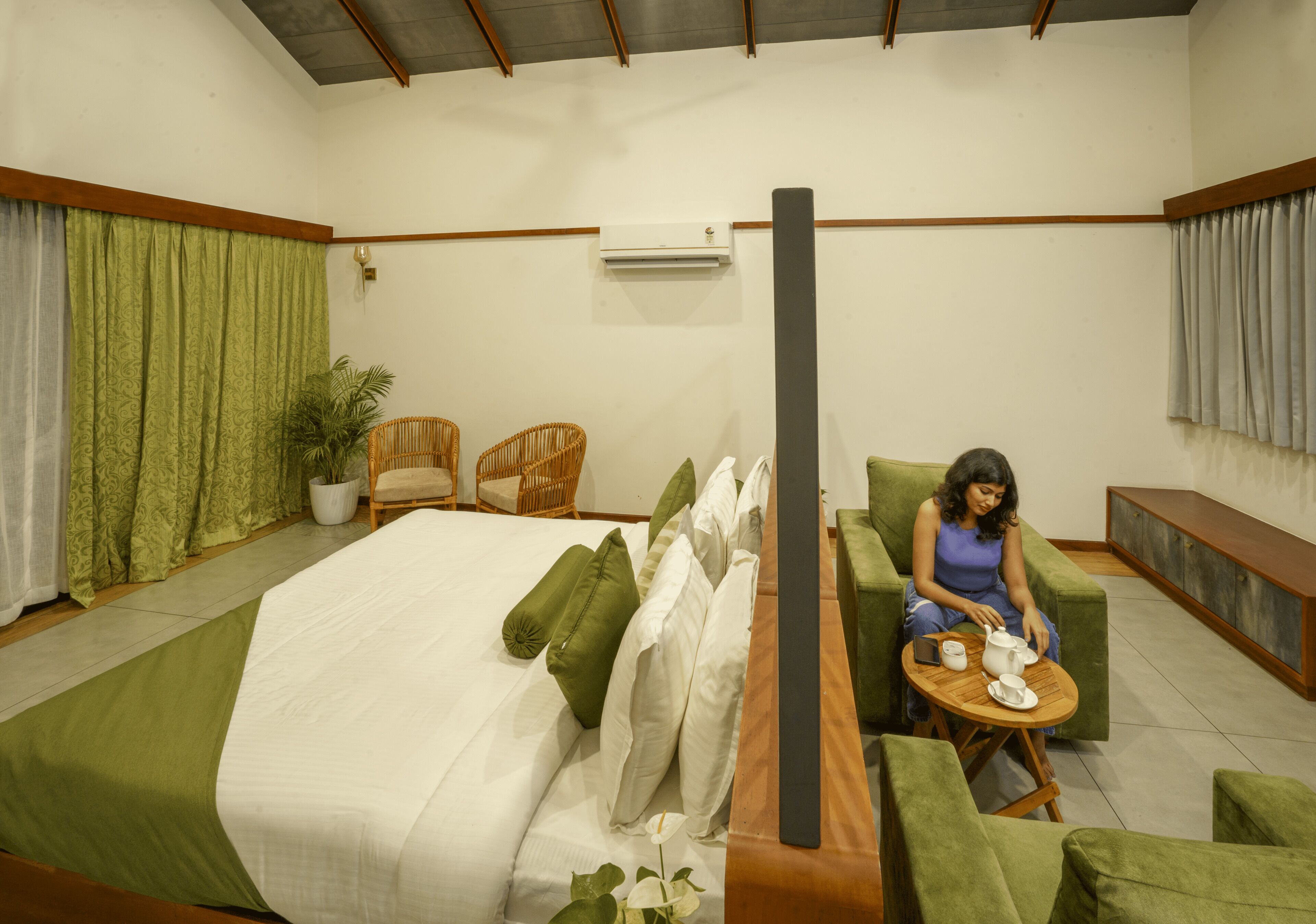 A room with a bed, green upholstered chairs, a small round table with a teapot, and a potted plant.