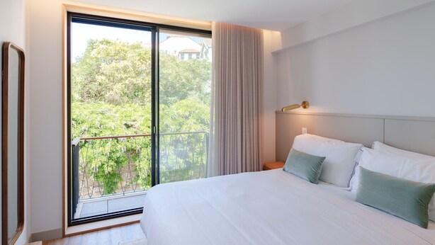 Habitación de hotel moderna con una cama grande, una mesita de noche, una lámpara y vistas a zonas verdes a través de una puerta corrediza de vidrio.