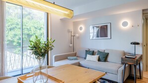Una sala de estar moderna con un sofá, una mesa de centro de madera, un florero con plantas y un ventanal con vistas a árboles.