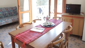 Un comedor con una mesa de madera, sillas, un mantel individual rojo, una cafetera, tazas y platos. A la derecha, hay un televisor sobre un mueble de madera.