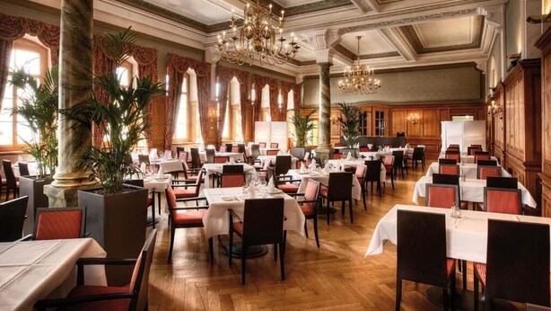 A formal dining room with multiple tables and chairs, large windows with curtains, and decorative columns. - Hotel Bellevue (Alpnach)