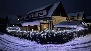 Ein mit Schnee bedecktes Haus mit einem gut beleuchteten Garten, einem sichtbaren Eingang und einer verschneiten Auffahrt.