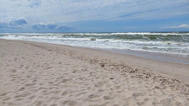 Ein sandiger Strand mit Fußabdrücken, die zum Wasser führen, ein klarer Himmel und Wellen, die an die Küste rollen.