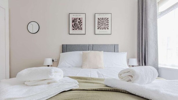 Ein Hotelzimmer mit einem ordentlich bezogenen Bett, zwei gerahmten Kunstwerken an der Wand, einem runden Spiegel und einem Fenster mit Vorhängen.