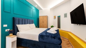Een moderne hotelkamer met een wand in teal, een groot bed met een donkerblauwe hoofdbord, wit beddengoed en een blauwe sprei. Er is een houten garderobe, een flatscreen-tv aan de muur en een klein nachtkastje met een lamp.