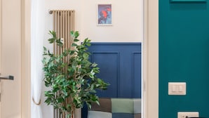 Een kamer met houten vloer, een blauwe deur, een radiator, een plant in een witte pot, een bed met een gestreept dekbed en een kleine tafel met een lamp.