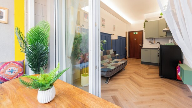 Een moderne kamer met een houten vloer, een bank, een kleine tafel met een plant, en een keukenhoek met kastjes en een koelkast.