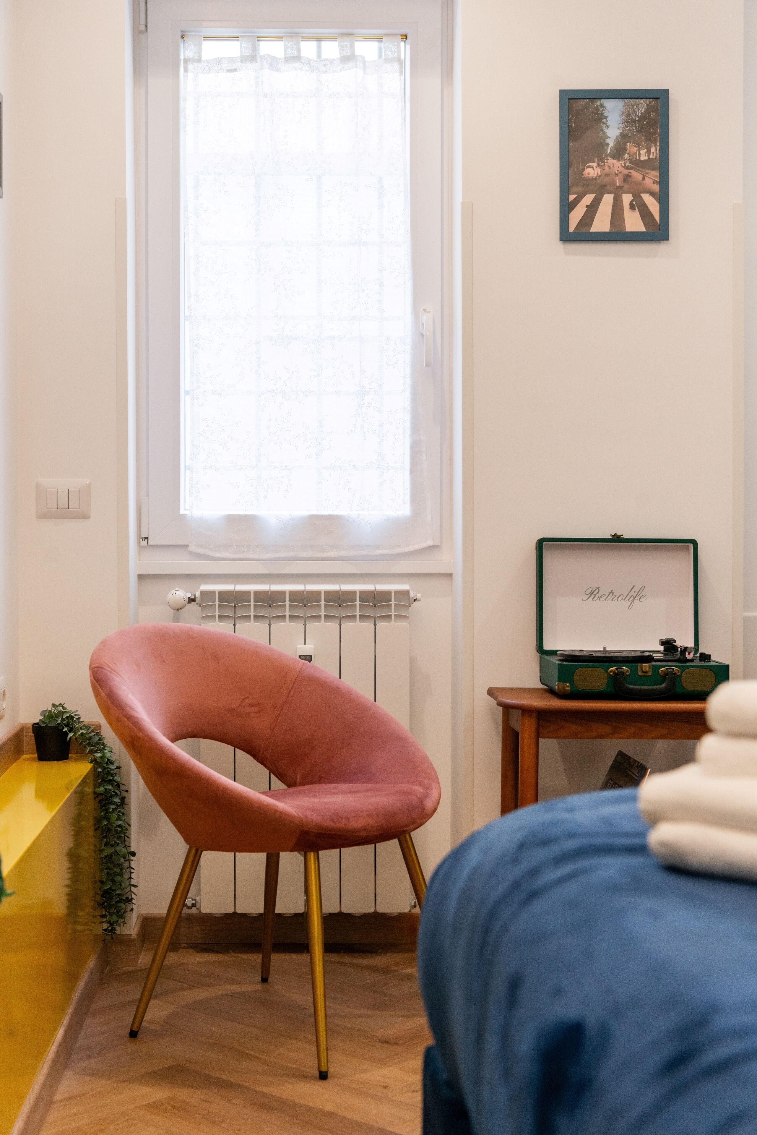 A room with a window, radiator, a pink chair, a green suitcase, and a framed picture on the wall.