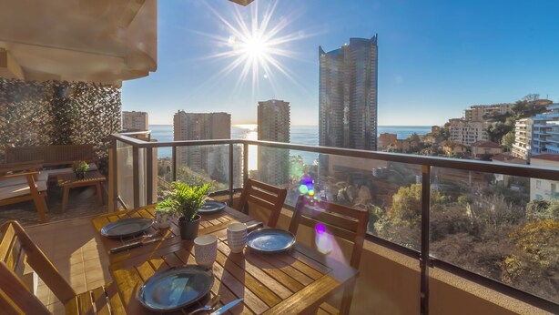 A balcony with a wooden table set for dining, overlooking a cityscape and the sea.