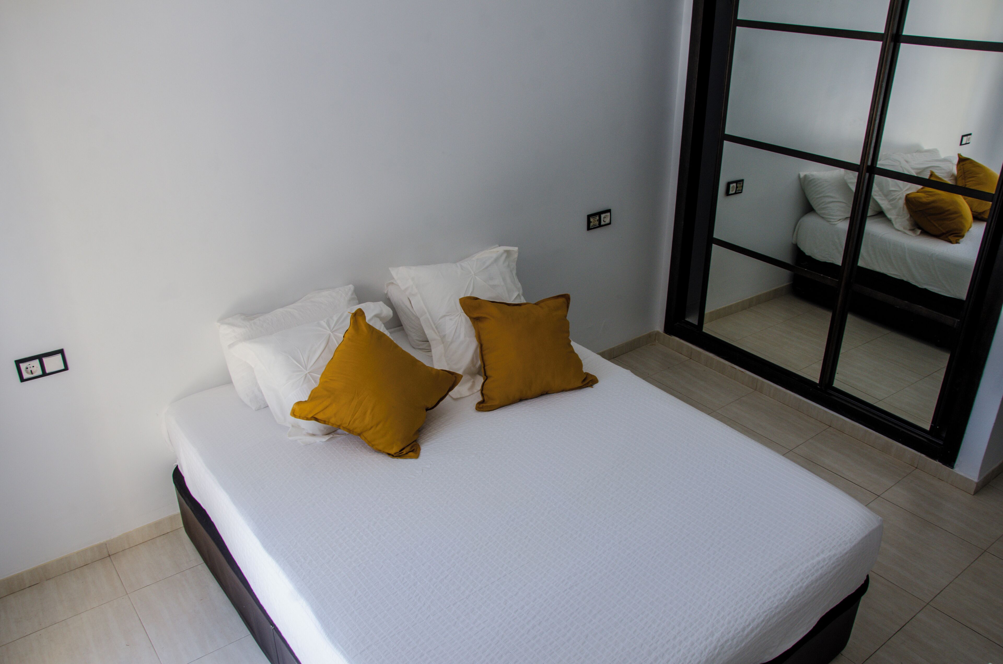 Una cama bien hecha con almohadas blancas y mostaza en una habitación con puerta corredera de vidrio que da a otra habitación.