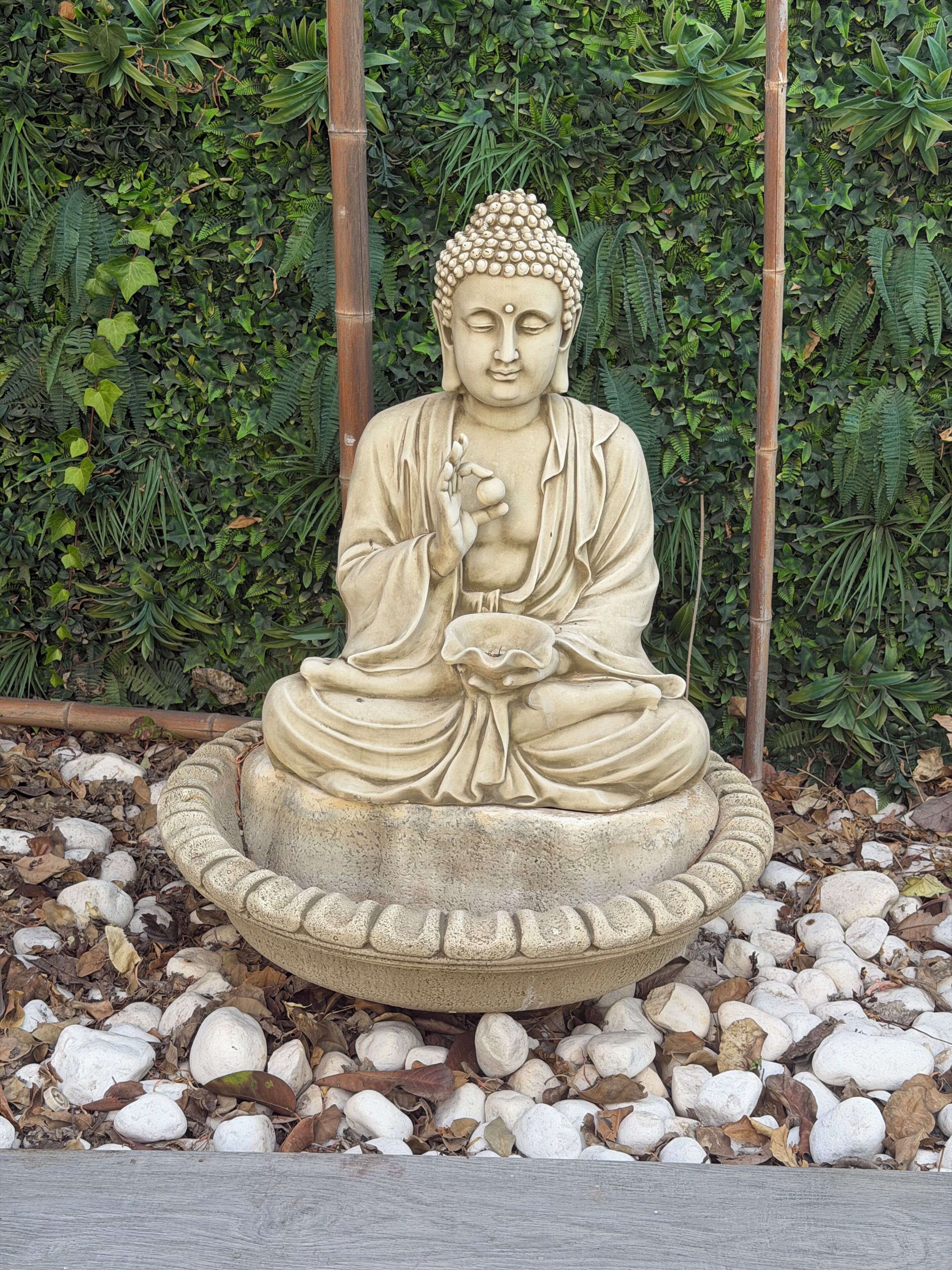 Une statue de Bouddha assis, placée dans un jardin, sur un socle en pierre, et entourée de feuillage et de rochers.