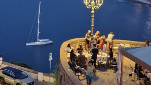 Un espace de restauration en bord de mer, avec des tables et des chaises, donnant sur une étendue d’eau où l’on aperçoit un voilier.