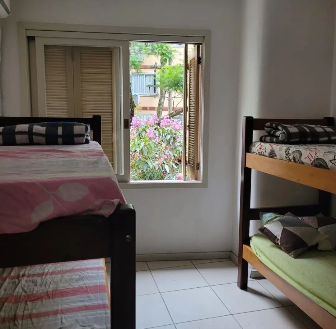 A dormitory room with two bunk beds, a window with shutters, and a view of a garden.