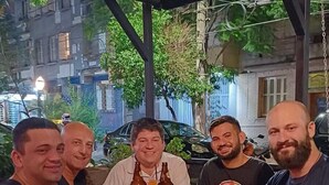 A group of people sitting at an outdoor table with drinks, under a covered area with a view of buildings and trees. - Man's Place Hostel Masculino (Porto Alegre)