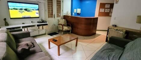 A living room with a sofa, a coffee table, and a TV on the wall. There is a reception desk with a blue wall and a door labeled "SAIDA".