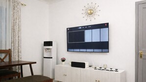 Una sala moderna con un sofá blanco, una mesa de comedor de madera, un mueble blanco con un televisor montado encima y un dispensador de agua.