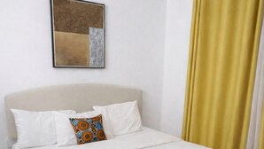 Una cama bien hecha con sábanas blancas, una toalla enrollada, un cabecero color beige y una sola almohada con un estampado colorido.