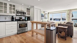 A modern kitchen with white cabinets, a built-in microwave, and an oven. There is a wooden table with a bench in the center. A refrigerator is on the right, and a sofa is in the living area with a view of the sea. - Oceanfront Getaway with Deck, Steps to Provincetown Fun (Provincetown)