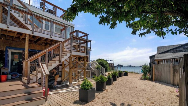 A wooden deck with a staircase leading to a building with large windows, a sandy beach in the background, and a view of the sea. - Oceanfront Getaway with Deck, Steps to Provincetown Fun (Provincetown)