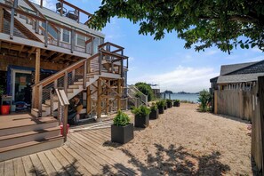 A wooden deck with a staircase leading to a building with large windows, a sandy beach in the background, and a view of the sea. - Oceanfront Getaway with Deck, Steps to Provincetown Fun (Provincetown)