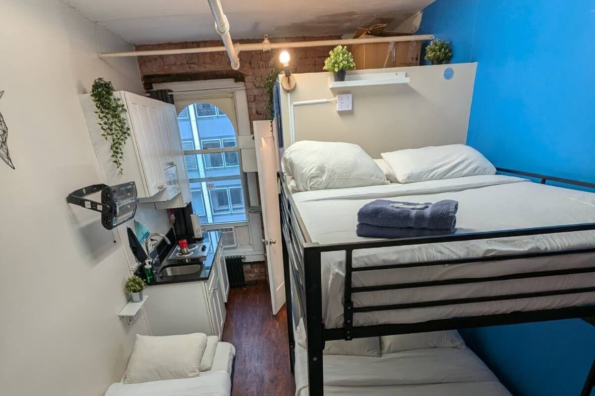 A dormitory room with bunk beds, a kitchenette, a radiator, and a window with a view.