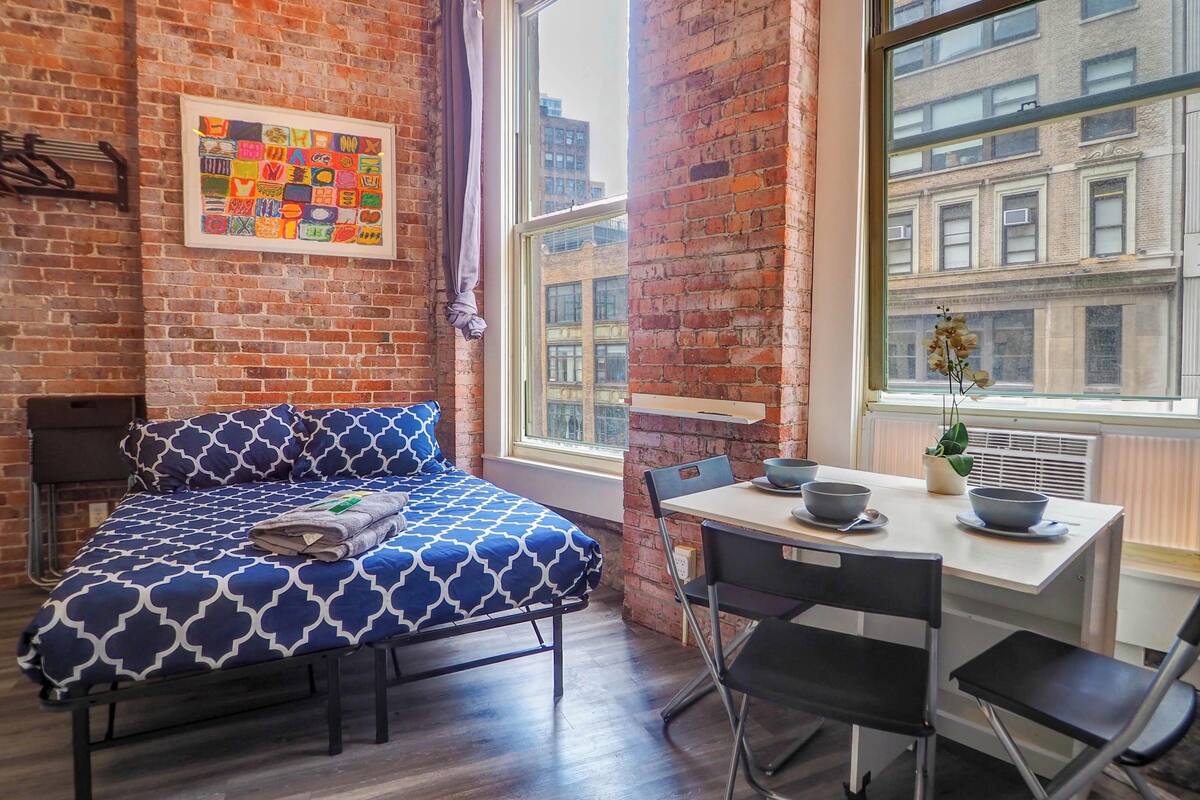 A room with a bed, a desk, and a window with a view of buildings.