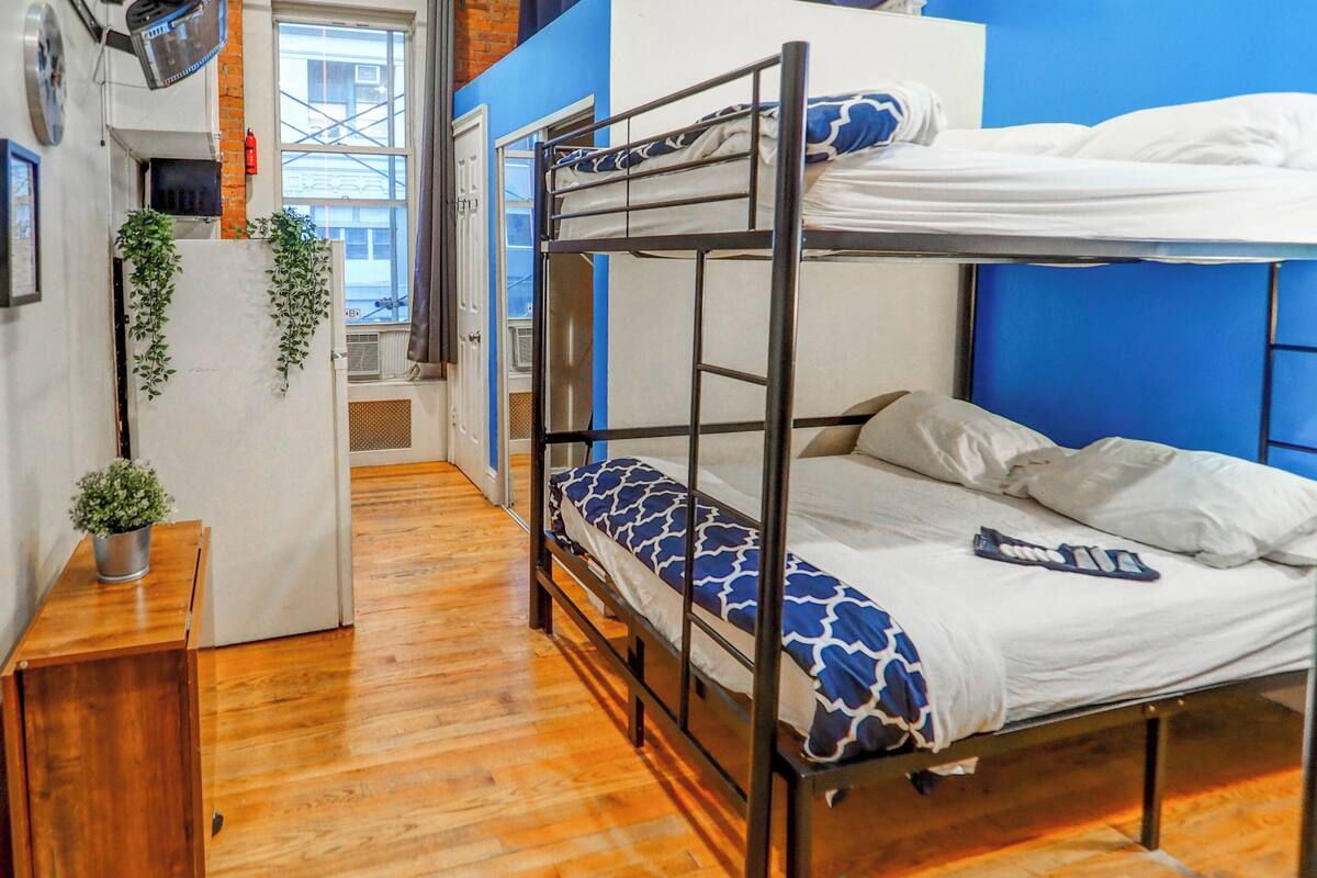 A bunk bed room with a wooden floor, a small desk with a plant, and a window with a view of buildings.