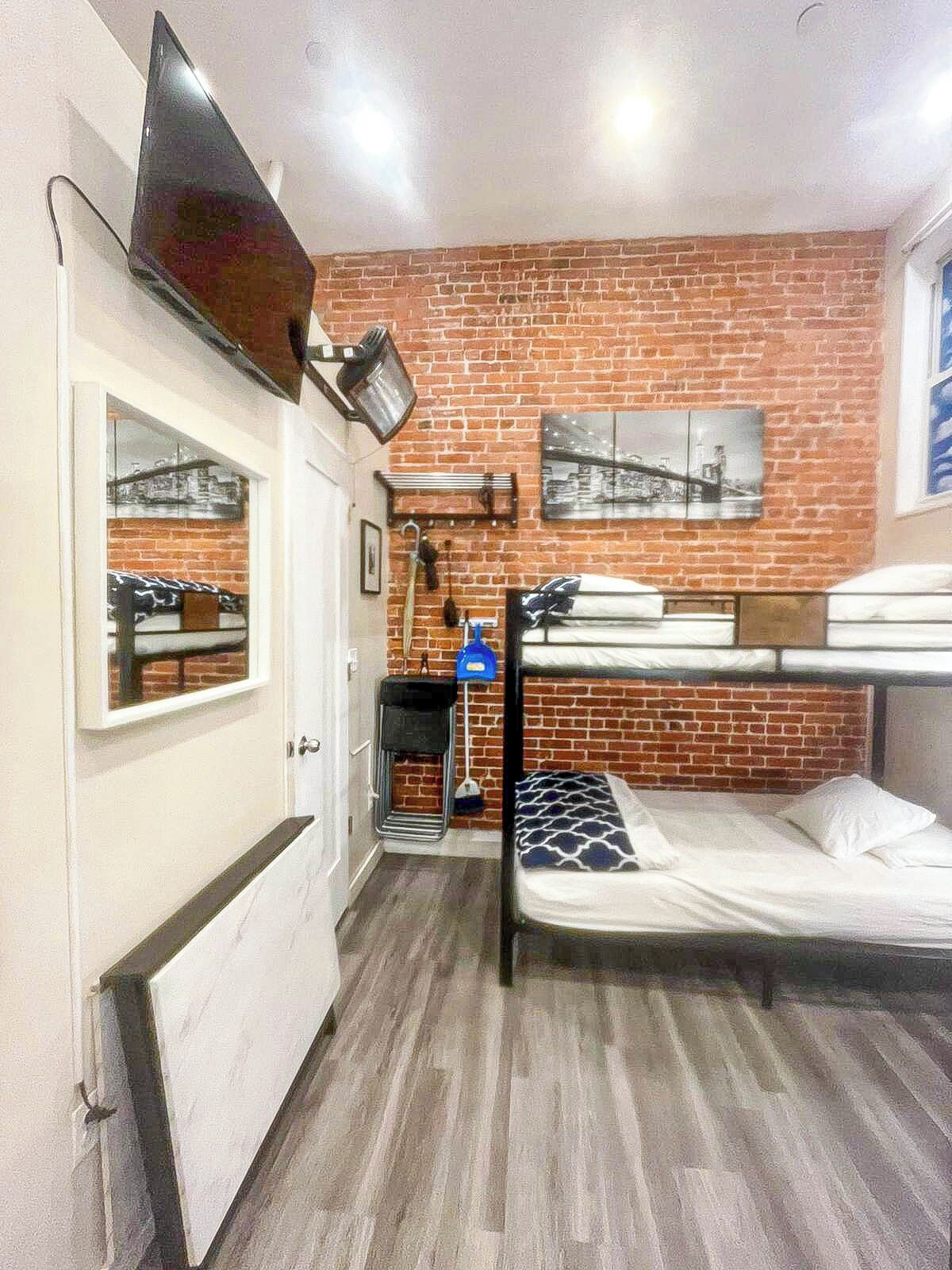 Una habitación pequeña con una cama individual superpuesta, un escritorio pequeño con una silla, un televisor montado en la pared y una pared de ladrillo con un cuadro enmarcado.