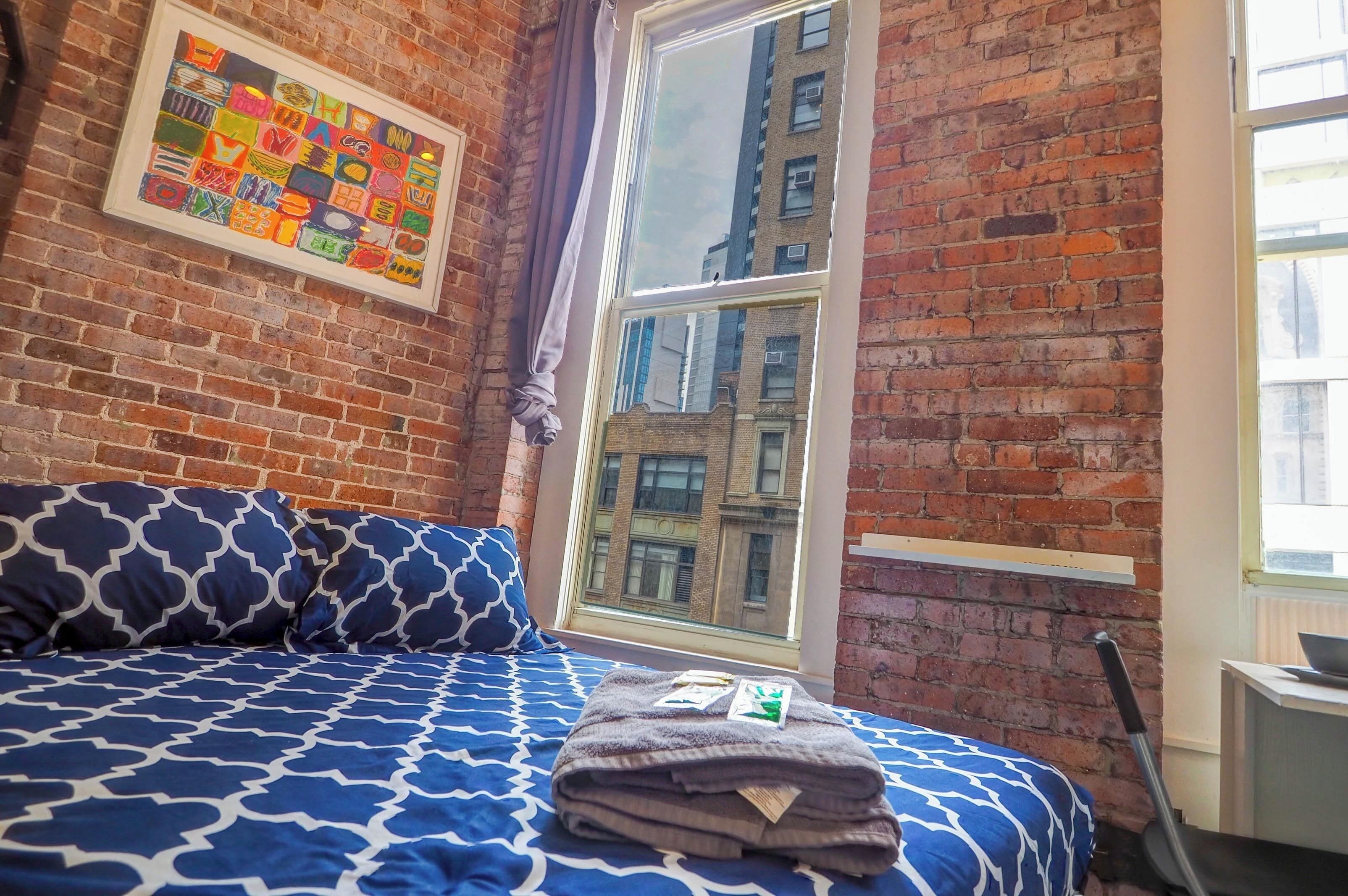 A room with a brick wall, a large window, a bed with patterned bedding, and a framed artwork on the wall.