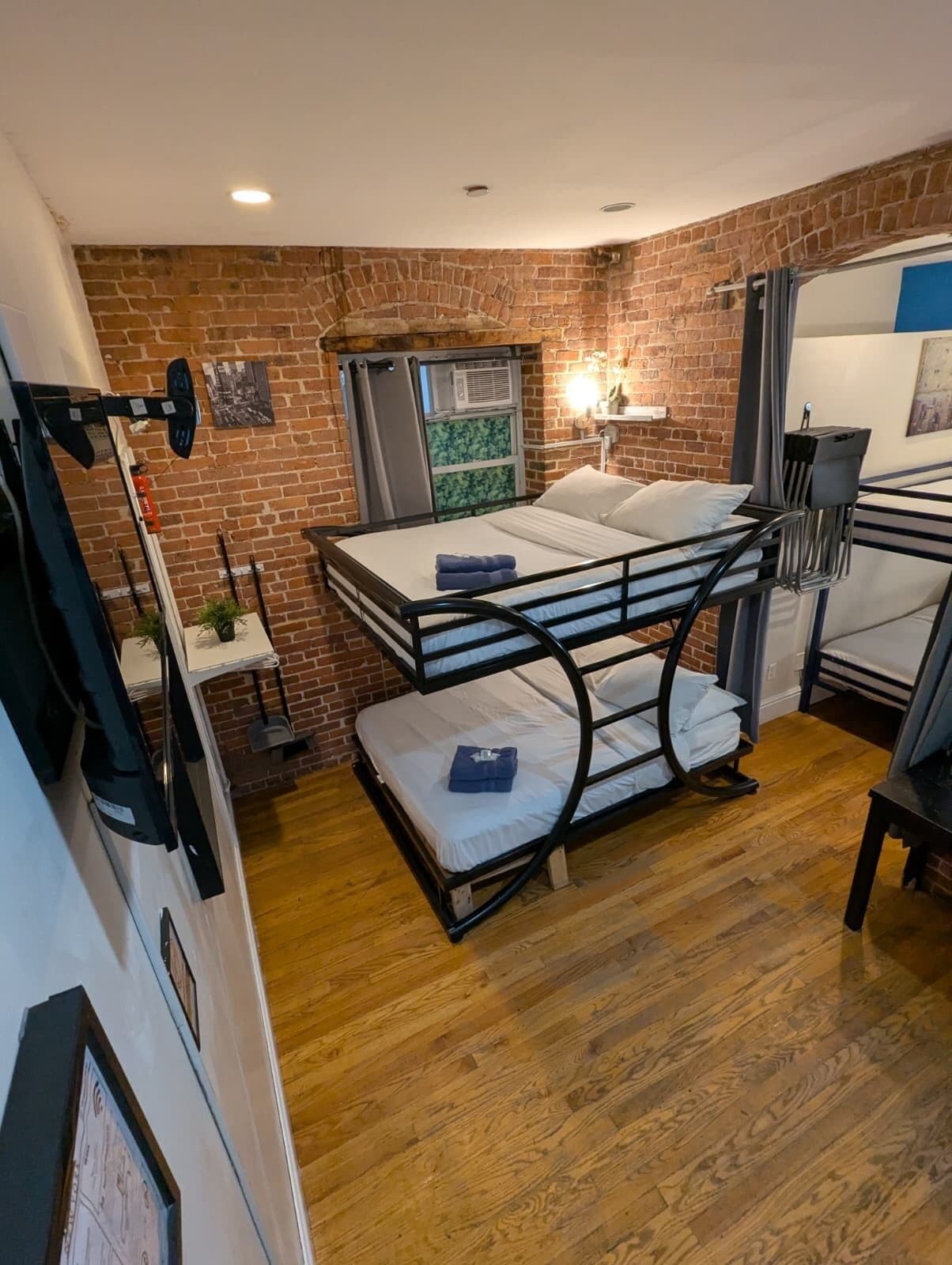 Ein kleines Zimmer mit Backsteinwand, Etagenbett, einem kleinen Tisch und einem Fenster mit Klimaanlage.