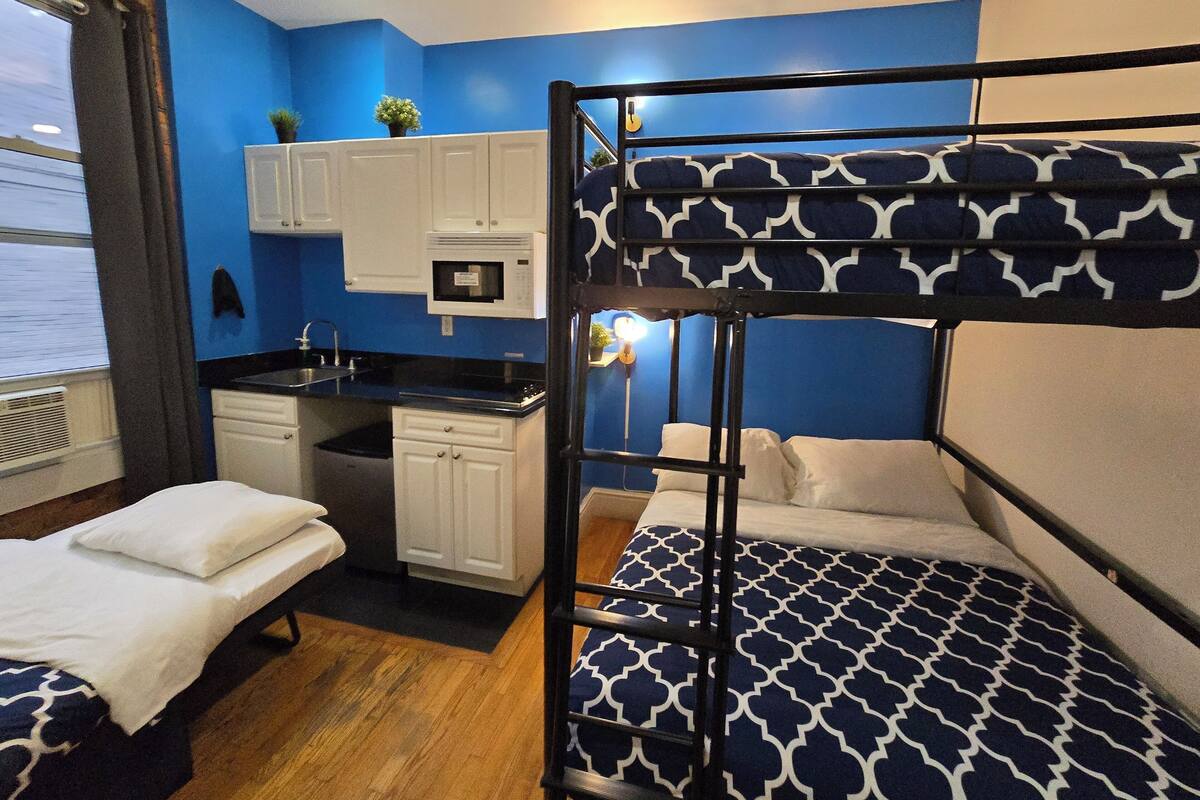 A compact room with bunk beds, a kitchenette, and a window with blinds.