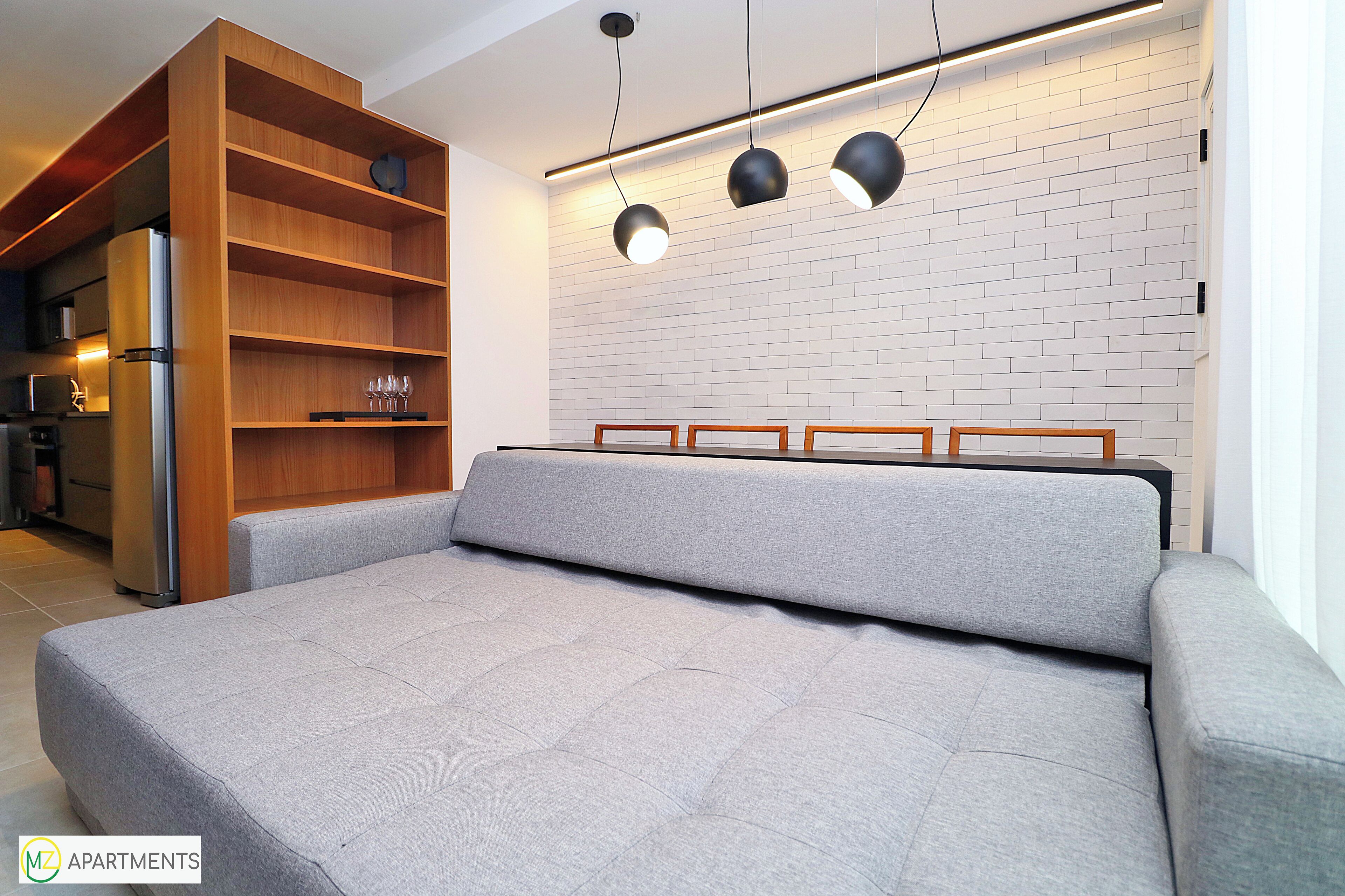 A modern living room with a grey sofa, wooden shelving unit, and a kitchen area with a refrigerator.