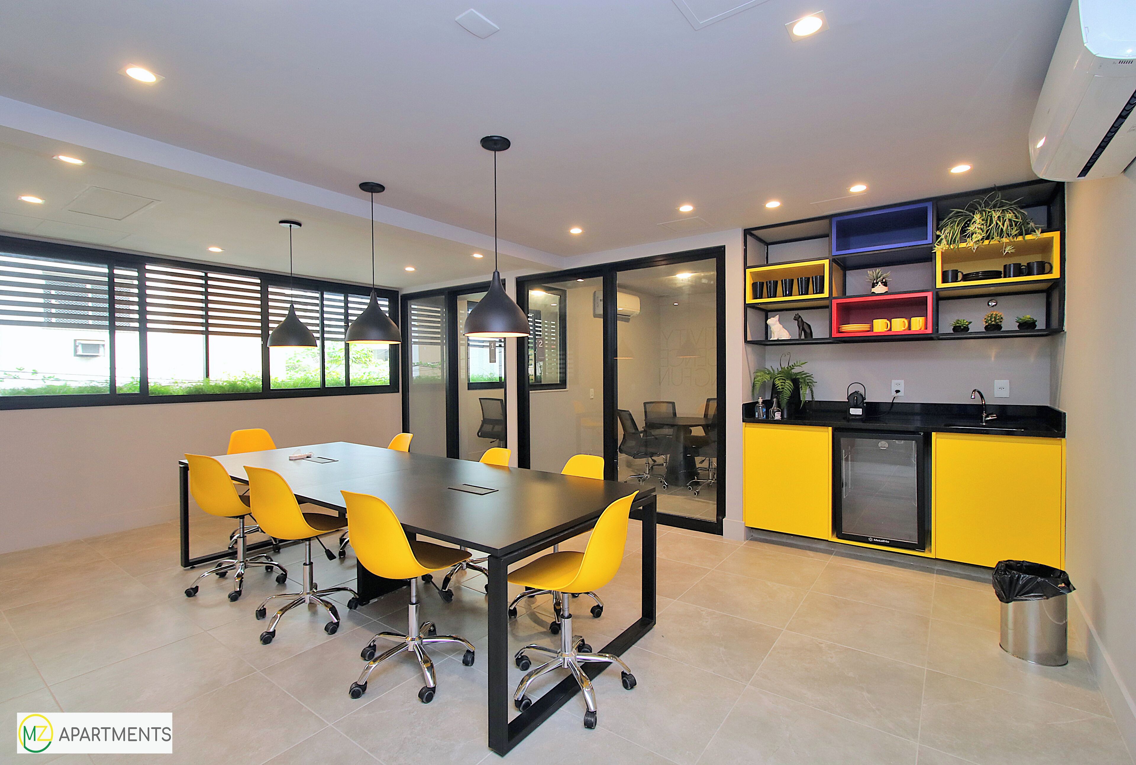 A modern meeting room with a large table, yellow chairs, pendant lighting, and a kitchen area with yellow cabinets and shelves.
