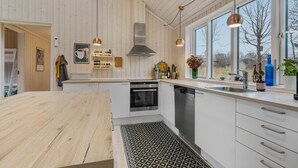 A modern kitchen with wooden walls, a central island, stainless steel appliances, and large windows offering a view of the outdoors. - Beautiful home in Fårevejle with WiFi (Fårevejle)
