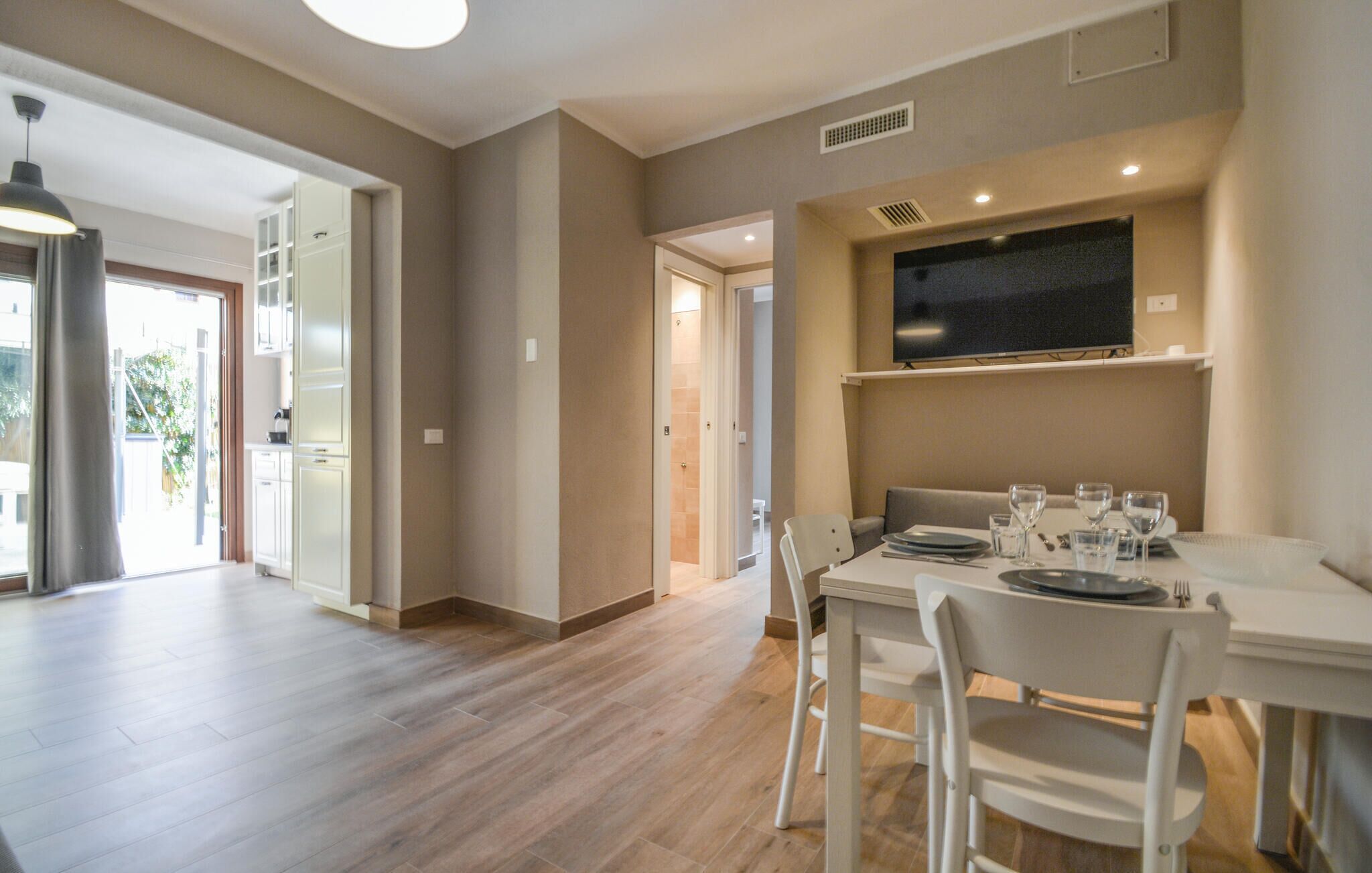 A dining area with a table set for four, a TV mounted above the table, and a hallway leading to a bathroom.