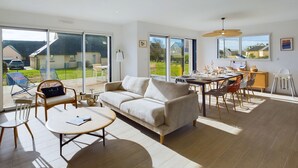 A spacious living room with a sofa, coffee table, and dining area. Large windows offer a view of the outdoors. - House for 10 - Garden - 600m from the beach (Penmarch)