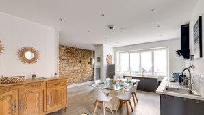 Une cuisine moderne avec une table à manger en verre, des chaises blanches, un meuble en bois et un mur en pierre.